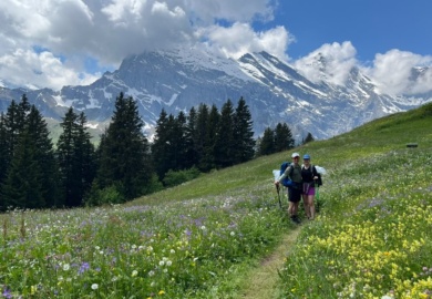 Pärchen Lea und Lucas beim Wandern auf dem Bärentrek auf Blumenwiese vor Gebirge in der Schweiz im Berner Oberland mit dabei ein Barfußschuh als Wanderschuh