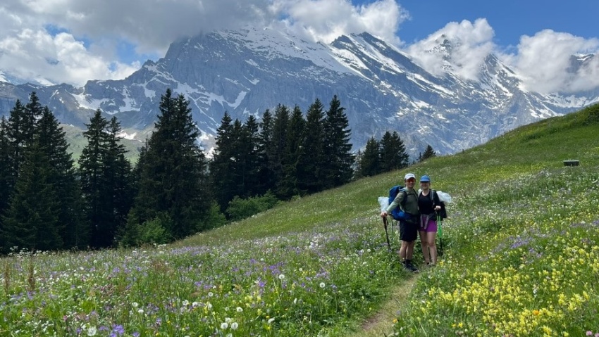 Pärchen Lea und Lucas beim Wandern auf dem Bärentrek auf Blumenwiese vor Gebirge in der Schweiz im Berner Oberland mit dabei ein Barfußschuh als Wanderschuh