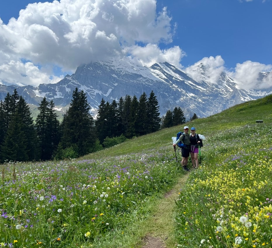 Pärchen Lea und Lucas beim Wandern auf dem Bärentrek auf Blumenwiese vor Gebirge in der Schweiz im Berner Oberland mit dabei ein Barfußschuh als Wanderschuh